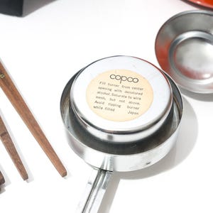 May include: A collection of vintage kitchen tools, including a metal burner with a label reading "copco" and instructions. Also includes wooden-handled utensils and a metal ladle. The items are arranged on a white surface.