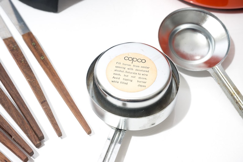 May include: A collection of vintage kitchen tools, including a metal burner with a label reading "copco" and instructions. Also includes wooden-handled utensils and a metal ladle. The items are arranged on a white surface.