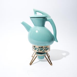 May include: A vintage turquoise ceramic teapot with a lid and a curved handle. It sits on a decorative metal stand with a gold finish and three legs. The teapot has a unique, rounded shape.