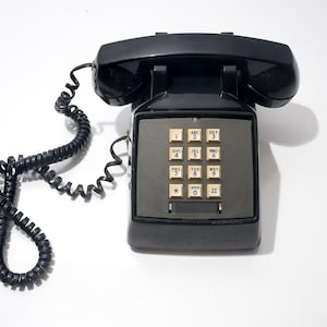Może przedstawiać: Czarny, zabytkowy telefon z słuchawką i zwiniętym kablem. Telefon posiada klawiaturę z beżowymi przyciskami i szarą płytą czołową. Klasyczny design, prawdopodobnie z połowy XX wieku.