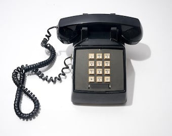 May include: A black vintage telephone with a handset and a coiled cord. The phone has a keypad with beige buttons and a gray faceplate. The phone is a classic design, likely from the mid-20th century.
