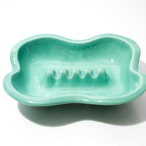 May include: A vintage, turquoise-colored ashtray with a unique, wavy shape. The ceramic ashtray features four cigarette rests and a glossy finish. A decorative piece for a home or office.
