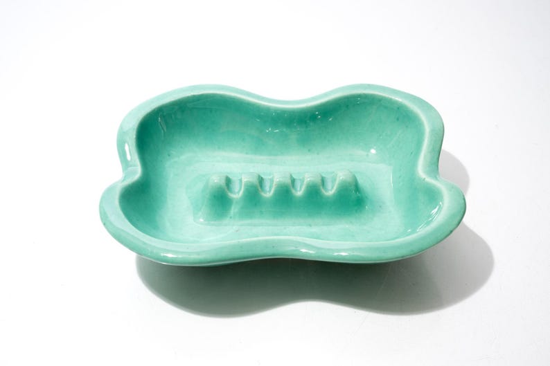 May include: A vintage, turquoise-colored ashtray with a unique, wavy shape. The ceramic ashtray features four cigarette rests and a glossy finish. A decorative piece for a home or office.