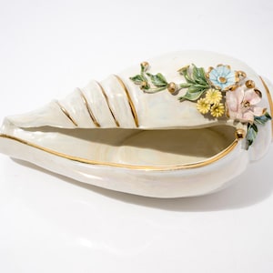 May include: A white ceramic conch shell bowl featuring a gold-trimmed rim and spiral ridges. The exterior is decorated with a cluster of sculpted, hand-painted flowers in shades of light blue, yellow, and pink, accompanied by green leaves.