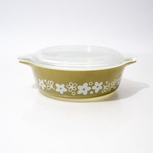 Puede incluir: Una fuente de horno redonda de vidrio color verde oliva con dos asas laterales integradas y una tapa de vidrio transparente. El exterior de la fuente presenta un patrón floral blanco repetitivo que consiste en varias formas de flores estilizadas y pequeños puntos.