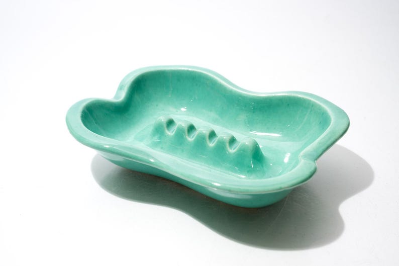May include: A vintage, turquoise ashtray with a unique, cloud-like shape. The ceramic ashtray features four cigarette rests and a glossy finish. A retro piece for home decor.