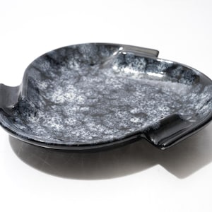 May include: A shallow, rounded ceramic dish featuring a glossy black and white mottled glaze. The dish has a smooth, curved rim with three distinct, raised rectangular notches spaced evenly around the perimeter, creating a unique silhouette.