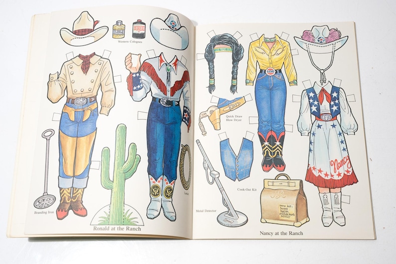 May include: A vintage paper doll book featuring cowboy and cowgirl outfits and accessories. The illustrations include hats, shirts, pants, boots, and a cactus. The text reads "Ronald at the Ranch" and "Nancy at the Ranch".
