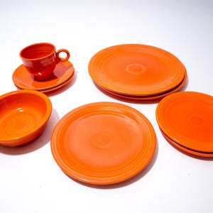 Pode incluir: Um conjunto de louça de cerâmica laranja, incluindo pratos, uma tigela, uma chávena e um pires. Os pratos estão empilhados, e a tigela e a chávena estão posicionadas perto dos pratos. O conjunto está disposto sobre uma superfície branca.