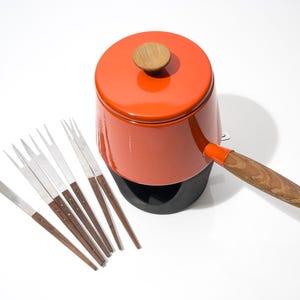 May include: An orange fondue pot with a wooden handle and lid, accompanied by six fondue forks. The pot sits on a black base. The forks have stainless steel tines and wooden handles.