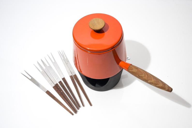 May include: An orange fondue pot with a wooden handle and lid, accompanied by six fondue forks. The pot sits on a black base. The forks have stainless steel tines and wooden handles.