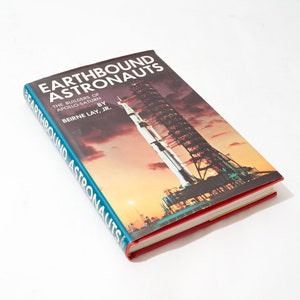 May include: A teal blue hardcover book titled "Earthbound Astronauts: The Builders of Apollo-Saturn" by  Berne Lay, Jr. The cover features a photograph of a rocket launch at night.