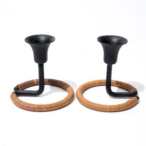 May include: Two black metal candle holders with a circular rattan base. The candle holders have a curved design, with a black cup for the candle and a rattan ring base. The candle holders are on a white background.