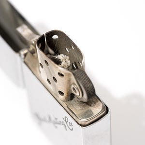 K&ouml;nnte beinhalten: Nahaufnahme eines silbernen Metallfeuerzeugs mit ge&ouml;ffneter Oberseite. Das Feuerzeug hat einen rechteckigen K&ouml;rper und einen Klappdeckel. Der obere Teil hat einen Docht und ein Z&uuml;ndrad. Das Feuerzeug befindet sich auf wei&szlig;em Hintergrund.
