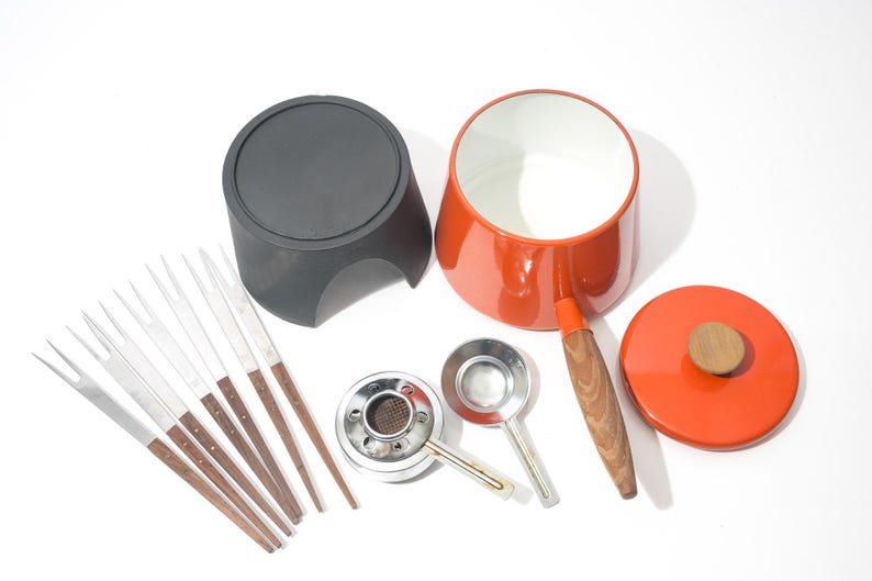 May include: A vintage fondue set with a red pot, black base, and lid with a wooden knob. Includes fondue forks, a burner, and two small ladles. The pot has a wooden handle. The set is designed for serving melted cheese or chocolate.