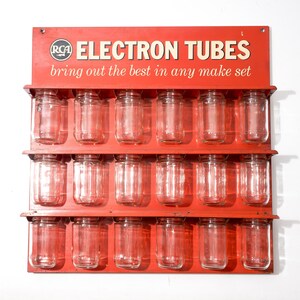 May include: A red metal wall-mounted display rack featuring the RCA logo and the text "ELECTRON TUBES bring out the best in any make set." The rack has three horizontal shelves holding a total of eighteen clear glass jars in three rows.