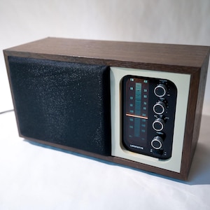 Peut inclure: Une radio AM/FM vintage en bois avec un haut-parleur noir et un cadran blanc avec un affichage numérique bleu et des boutons de réglage. La radio est étiquetée "Lafayette".