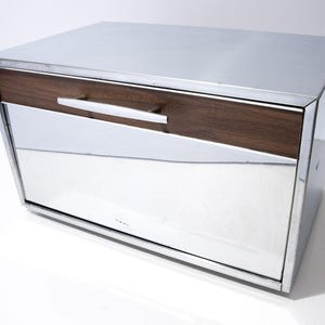 Könnte beinhalten: Ein rechteckiger Brotkasten im Mid-Century-Modern-Stil mit Edelstahl-Außenseite und einer Schublade aus walnussfarbenem Holz. Die Schublade hat einen silbernen Griff. Die Front ist verspiegelt. Auf weißer Oberfläche.