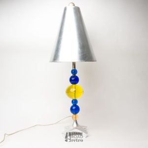 May include: A silver table lamp with a conical lampshade and a base made of blue and yellow glass spheres.