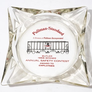 Può includere: Posacenere in vetro trasparente con un design a stella. Il centro presenta il testo "Pullman-Standard" e un disegno di un vagone ferroviario. Il testo recita "Butler 1966-Winner Annual Safety Contest Award to Employees."