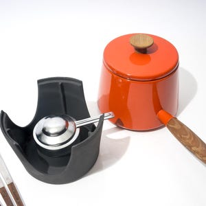 May include: An orange enamel saucepan with a wooden handle and lid, a black scoop holder with a stainless steel scoop, and a set of fondue forks. The kitchenware is set against a white background.
