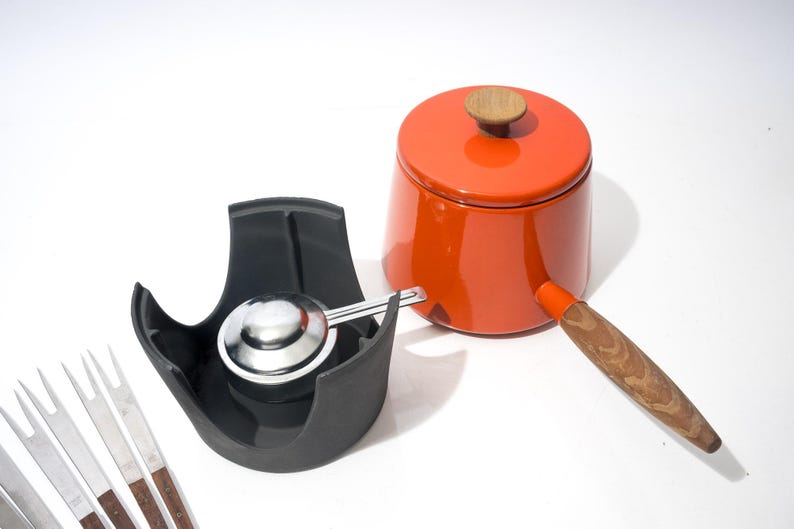 May include: An orange enamel saucepan with a wooden handle and lid, a black scoop holder with a stainless steel scoop, and a set of fondue forks. The kitchenware is set against a white background.