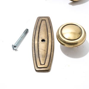 May include: A set of brass-toned cabinet hardware, including a rectangular pull with a central hole, a round knob, and a silver screw. The hardware has a vintage aesthetic.