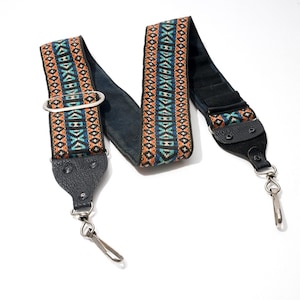Peut inclure: Une bandoulière de sac de remplacement avec une bande en tissu tissé ornée d'un motif géométrique orange, bleu sarcelle et bleu foncé. Elle comprend des embouts en cuir noir, des mousquetons pivotants argentés et une boucle de réglage argentée.