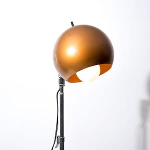 Puede incluir: Una lámpara esférica vintage de color cobre con una luz brillante que emana de la parte inferior abierta. La lámpara está montada sobre un soporte ajustable negro y plateado. El fondo es blanco liso.