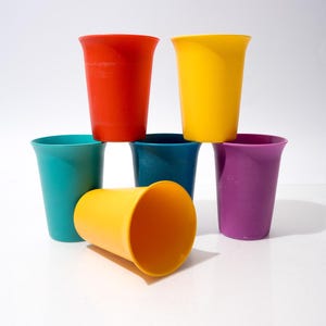 Könnte beinhalten: Eine Sammlung bunter Plastikbecher in Rot, Gelb, Türkis, Dunkelblau und Lila. Ein gelber Becher liegt auf der Seite, während die anderen gestapelt sind. Die Becher haben ein einfaches, abgerundetes Design.