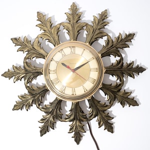 May include: A vintage wall clock with a gold-toned face and Roman numerals. The clock is surrounded by an ornate, gold-colored, leaf-shaped frame. The clock hands are black, with a red second hand. A power cord extends from the bottom.