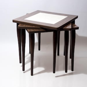 Puede incluir: Un juego de tres mesas nido con marcos de madera de color marrón oscuro e incrustaciones de azulejos blancos cuadrados en las superficies. Cada mesa se apoya sobre cuatro patas cónicas y están apiladas para mostrar sus alturas variables.