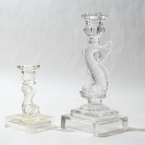 May include: Two clear glass candle holders. The larger one features a dolphin design, while the smaller one has a simpler design. Both have square bases and are suitable for standard-sized candles. The glass is clear and reflects light.