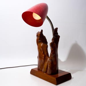 May include: A table lamp featuring a polished natural wood base and a vertical driftwood-style support. A flexible metal neck extends from the wood, holding a conical red lampshade with a single light bulb visible inside the shade.