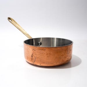 May include: A small copper saucepan with a brass handle. The interior is stainless steel. The pan is round and sits on a white surface. The handle is angled upwards.