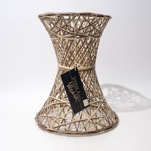 May include: Woven hourglass-shaped side table. The table is constructed from light and dark brown woven material. A black tag with white text hangs from the center. The table has a wide base and top, with a narrower center.