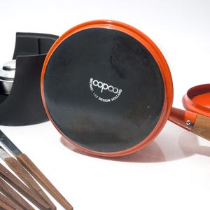 May include: An orange fondue pot with a wooden handle and a black base, accompanied by a black stand and several fondue forks. The pot is marked "copco" and "MICHAEL LAX DESIGN - HOLLAND".