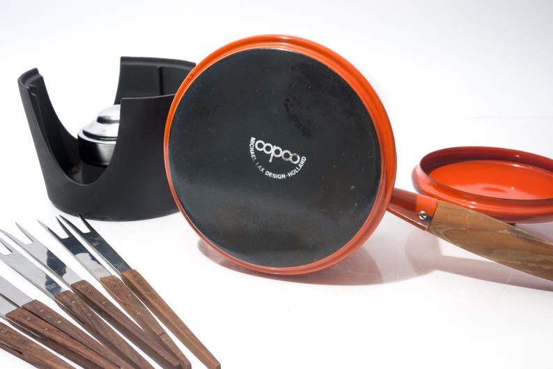 May include: An orange fondue pot with a wooden handle and a black base, accompanied by a black stand and several fondue forks. The pot is marked "copco" and "MICHAEL LAX DESIGN - HOLLAND".