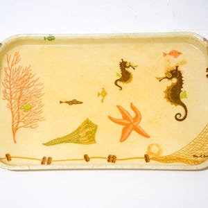 Può includere: Vassoio rettangolare con sfondo color crema e design a tema marino. Il vassoio presenta illustrazioni di cavallucci marini, pesci, una stella marina e coralli. Il bordo ha un motivo a corda decorativo.