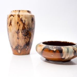 Puede incluir: Dos piezas decorativas de cerámica. Un jarrón alto y un cuenco bajo y redondo. Ambos tienen un patrón moteado en tonos marrón, beige y negro. El cuenco tiene un interior marrón oscuro. Las piezas están sobre una superficie blanca.