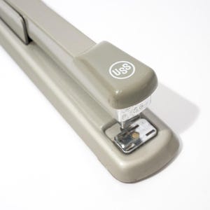 May include: A light gray stapler with the text "USS" in a white circle on the top. The stapler is on a white surface. The stapler is a common office supply used for fastening sheets of paper together.