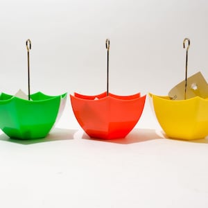 Pode incluir: Três vasos com forma de guarda-chuva coloridos, cada um com um gancho de metal. Os vasos são verde, vermelho e amarelo.