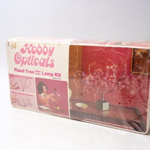 May include: A vintage "Hobby Opticals Floral Tree Fiber Optic Lamp Kit" box. The box features pink text and illustrations of the lamp and assembly instructions. The lamp is a decorative tree with fiber optic lights. The box is wrapped in clear plastic.