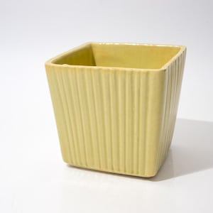 May include: A square, pale yellow ceramic planter with vertical ribbed detailing. The planter has a slightly tapered shape and a smooth, unadorned interior. The design is simple and modern, suitable for various plants.