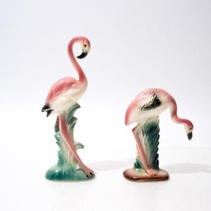 Pode incluir: Duas estatuetas de flamingo em cerâmica com plumagem rosa e branca e pontas das asas pretas. Um flamingo está de pé com o pescoço curvado, enquanto o outro é retratado numa postura voltada para baixo sobre bases verdes texturizadas.