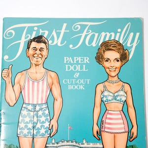 May include: A vintage "First Family" paper doll and cut-out book. The cover features illustrations of a man in a striped tank top and star-patterned shorts, and a woman in a star-patterned bra and striped skirt. The background is light blue.