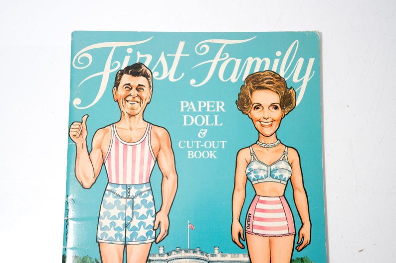 May include: A vintage "First Family" paper doll and cut-out book. The cover features illustrations of a man in a striped tank top and star-patterned shorts, and a woman in a star-patterned bra and striped skirt. The background is light blue.