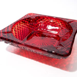 May include: A square, deep red glass ashtray featuring a faceted, diamond-cut pattern on the exterior and a sunburst design on the interior base. The edges are thick and translucent, catching the light to highlight the vibrant crimson color.