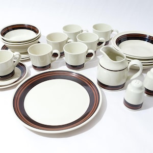 Peut inclure: Un ensemble de vaisselle en céramique blanche avec des rayures brunes. L'ensemble comprend des tasses, des soucoupes, un pichet, un bol et des assiettes.