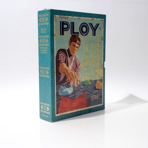 May include: Vintage board game box titled "PLOY" with a teal cover and spine. The front features an illustration of a man playing the game. The box has a retro design with text and graphics in blue, red, and yellow.